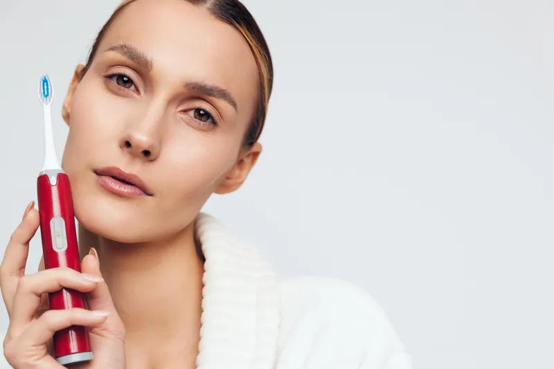Young woman holds electric toothbrush close to her face with gentle expression wearing white sweater isolated on plain light background for dental care concept.