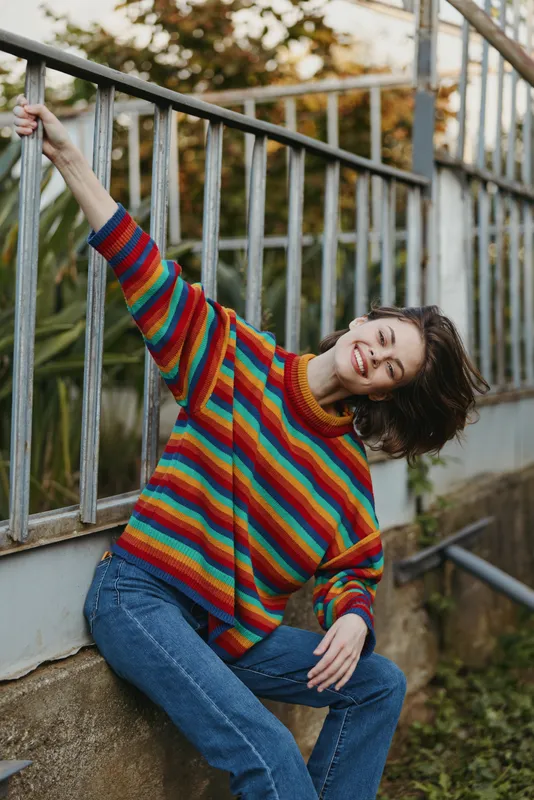 Woman smile rainbow sweater leaning on railing outdoor, casual portrait in jeans with joyful expression and relaxed pose near urban fence and greenery.