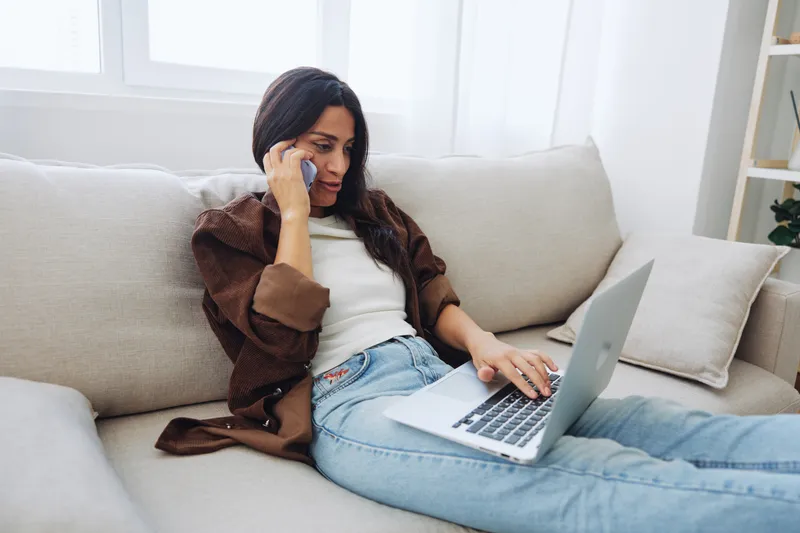 Женщина-блогер лежит дома на диване с телефоном, разговаривает по нему, рядом с ней ноутбук, она работает как фрилансер онлайн, делает селфи