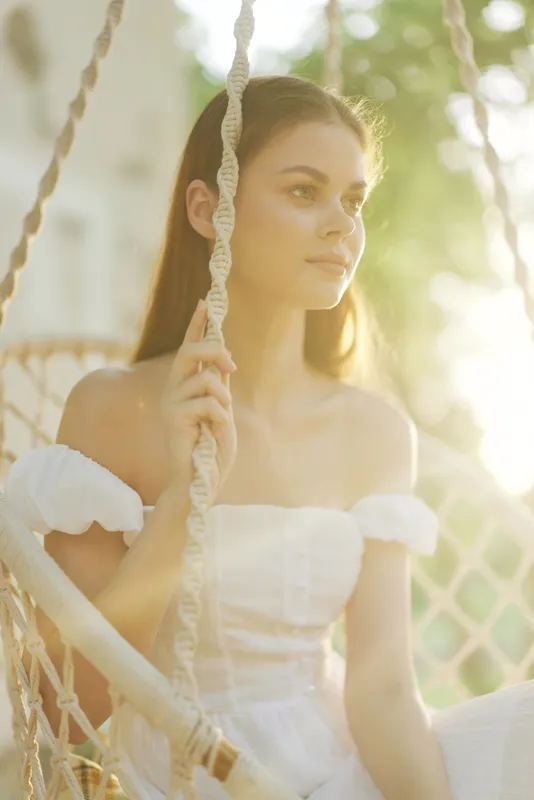 Serene young woman in white dress, basking in golden sunlight while sitting on a swing, evoking feelings of peace, summer, and tranquility