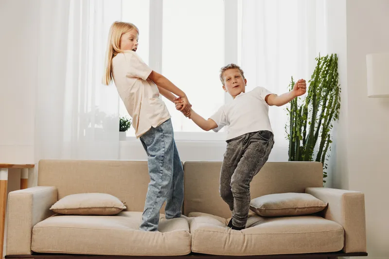 Two children holding hands and balancing on a beige sofa in a bright living room. Playful moment captures joy, friendship, and indoor activity with casual clothing and natural light.