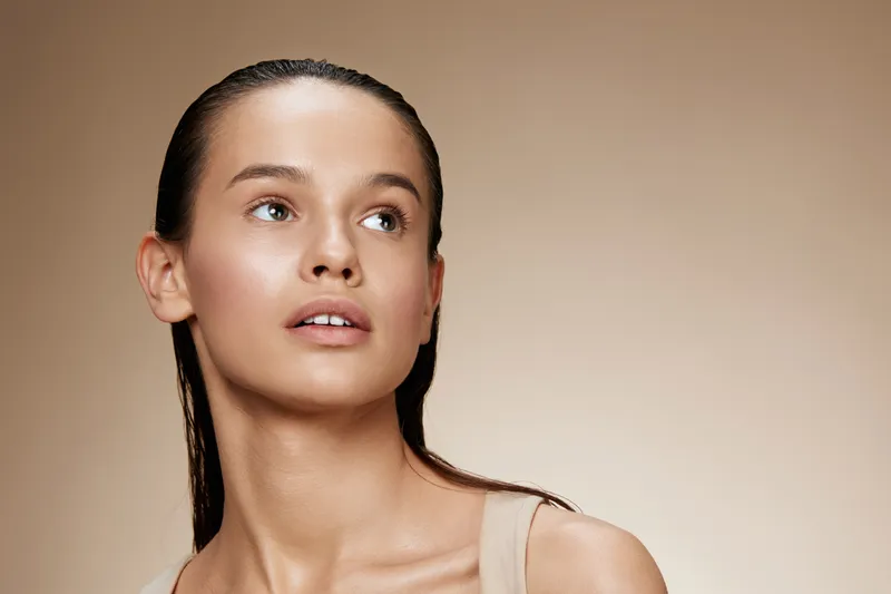 Portrait of a young woman with wet hair, showcasing natural beauty and radiance against a minimalistic beige background, evoking a fresh and serene atmosphere