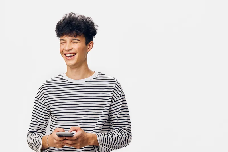 young man laughing holding smartphone in striped shirt isolated on white background, casual style, happy emotion, relaxed pose, modern technology