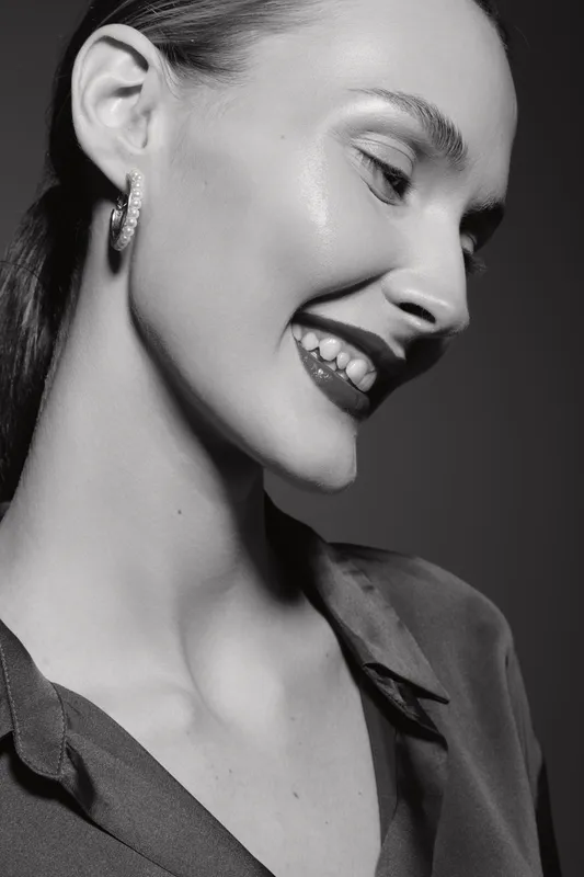Smiling Caucasian woman with fair skin and dark hair wearing hoop earrings and a shirt collar. Portrait in black and white with soft lighting and natural makeup. Concept of happiness and elegance.