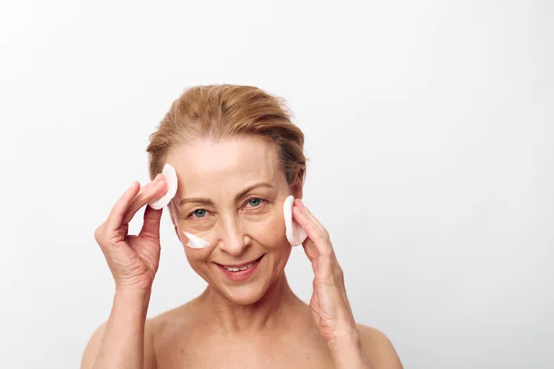 Mature woman with short hair gently applies cotton pads on her face, smiling softly against white background. Concept for skincare, beauty routine, and natural aging care.