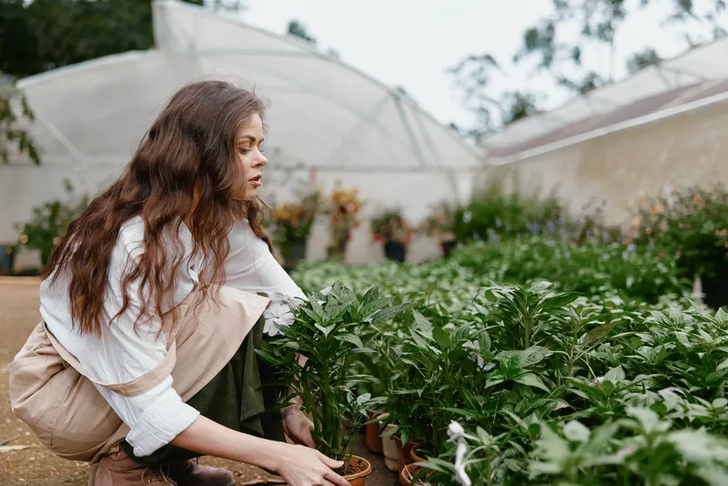 Young woman tending to plants in a greenhouse, showcasing dedication to gardening and nature, with a tranquil expression and casual attire in earthy tones
