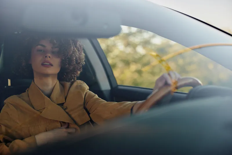 Woman driving a car with warm retro film color, wearing a stylish coat, sunlight on her face, candid lifestyle moment inside the vehicle, window view and detailed dashboard