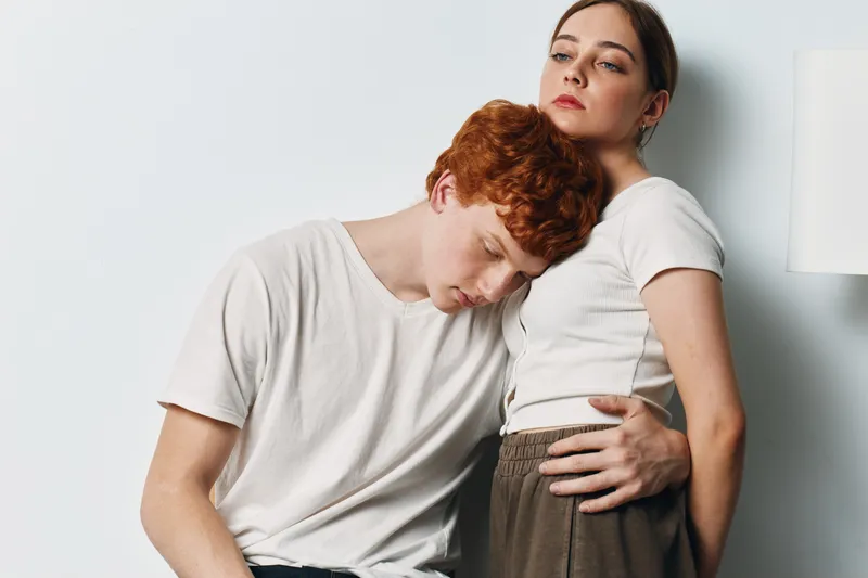 Young couple posing together with man leaning on woman shoulder for support, close relationship and intimacy in casual white outfits at home with soft natural light.