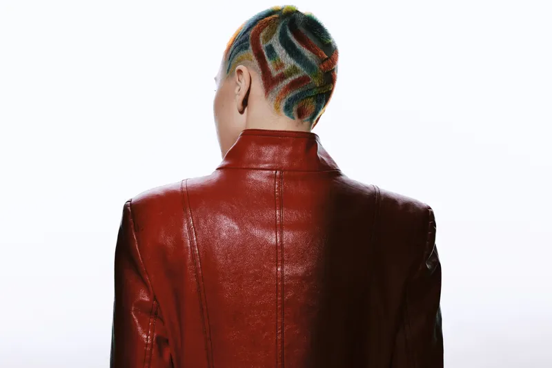 Woman with colorful shaved hairstyle wearing a glossy red leather jacket, viewed from behind against a bright white background, showcasing edgy modern fashion style.