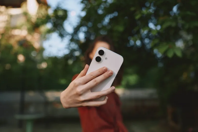 Smiling woman holding a smartphone for a casual outdoor selfie. Close-up with a blurred park background. Concept of lifestyle, wellness, and modern communication. Warm natural light creates a