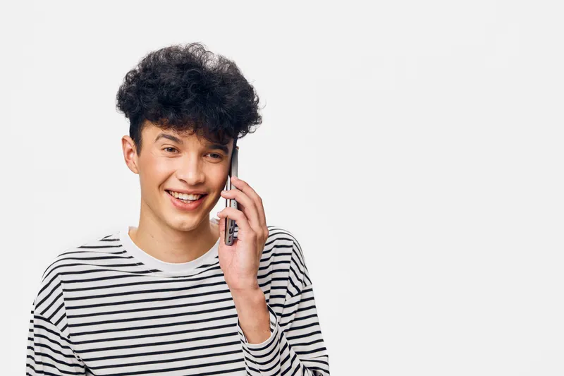 smiling man talking on smartphone in casual striped shirt standing against white background, youthful and friendly expression, modern communication concept.