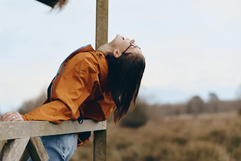 Woman lifestyle outdoors, leaning on wooden fence, wearing orange jacket and sunglasses, calm expression, warm sunlight, blurred rural field background, gentle breeze, candid photo