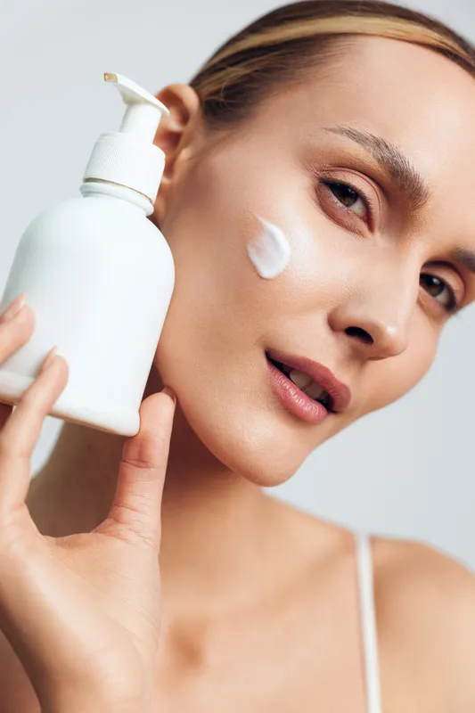 Young woman applying moisturizer on her cheek, holding white lotion bottle, natural skin care routine, clean background, fresh and healthy complexion, close up portrait