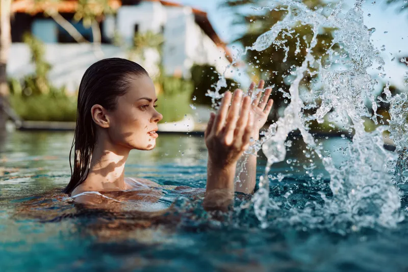 Young woman in swimming pool splashing water with hands, relaxed expression, natural light, outdoor leisure activity, summer lifestyle moment, clear water splash