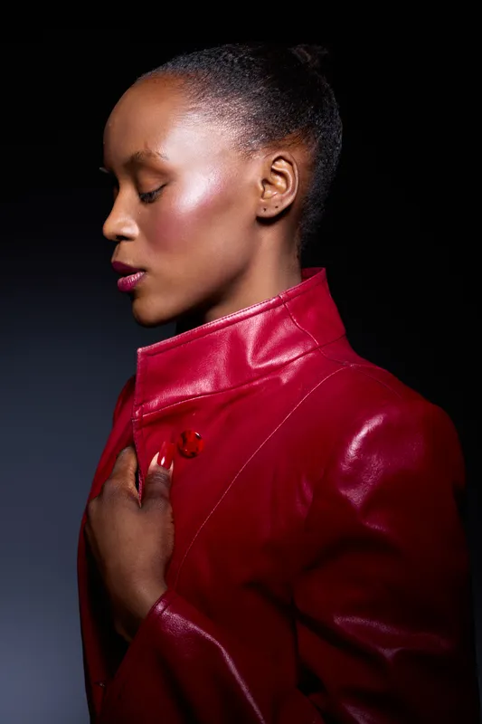 Profile of a young African woman with dark skin wearing a red leather jacket. She poses with closed eyes and a calm expression against a dark background. Concept of fashion and elegance.