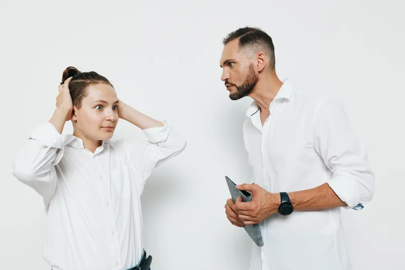 A professional man stands with a calm, confident gaze as a colleague adjusts her hair, against a plain white background. The moment highlights business focus, teamwork, and poise.