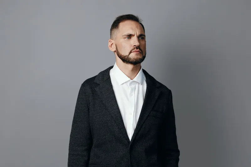 A professional businessman stands against a solid colored backdrop, showing determined confidence, calm focus, and poised energy in a modern work setting.