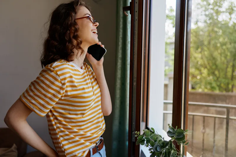 A casual woman in a striped tee stands by a sunlit window, talking on a smartphone, smiling softly. A home setting emphasizes everyday communication and relaxed, intimate moments.