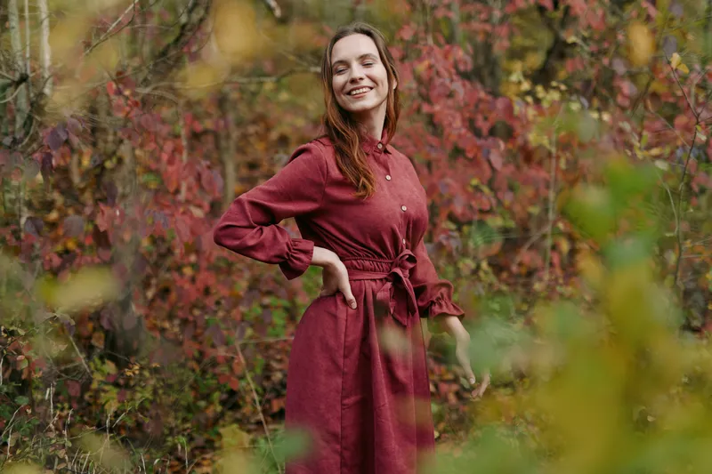 A candid portrait of a woman in a burgundy dress standing among autumn foliage, radiating warmth, confidence, and authenticity through soft natural light.