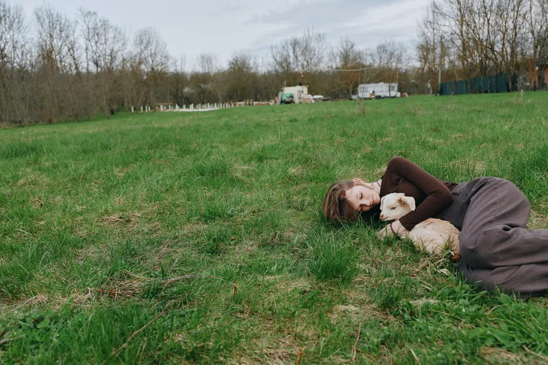 A woman lies on the grass in a park, cuddling a dog as soft sunlight washes the scene, conveying relaxation, companionship, lifestyle, park, tranquil, candid moments outdoors.