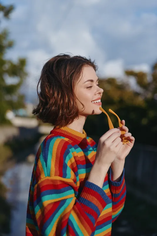Young woman in a rainbow sweater holds yellow sunglasses, smiling profile with short brown hair in an outdoor portrait. Casual outfit, relaxed mood, golden hour lifestyle and fashion vibe.