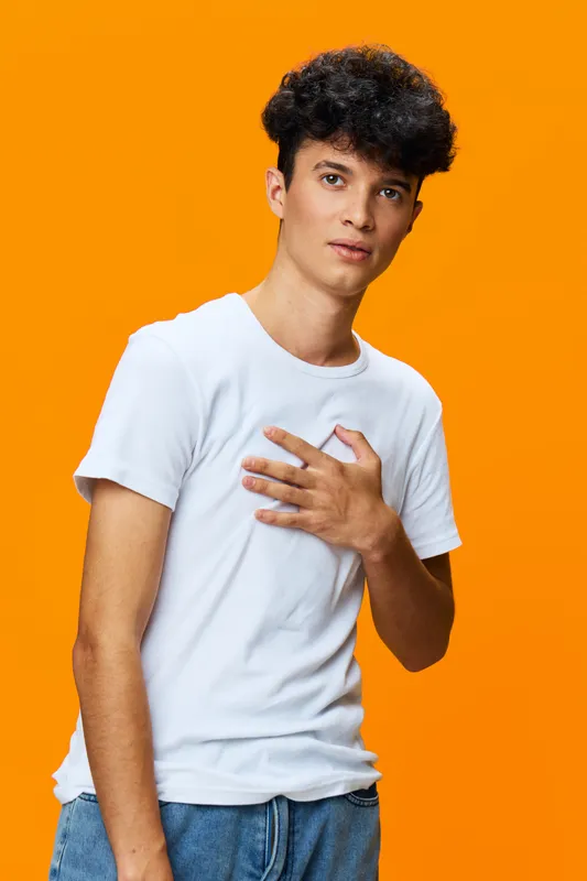 young man in white t-shirt expressing concern with hand on chest against vibrant orange background, casual style, thoughtful mood, isolated portrait