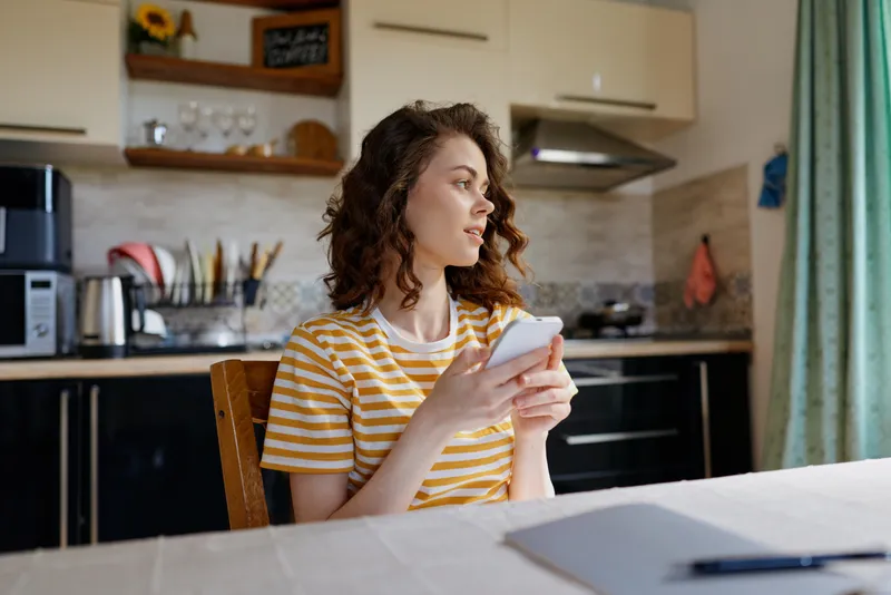 Casual portrait of a woman in a striped t shirt indoors at home, using a smartphone in a bright kitchen, relaxed lifestyle vibe, natural light, everyday moment captured with warmth. glow.