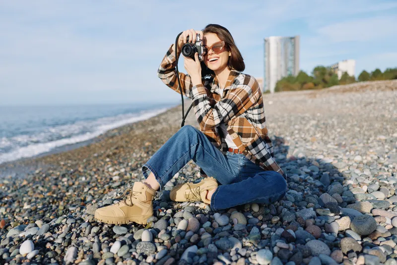 Casual woman with camera on a rocky beach, capturing moments by the sea in daylight. Outdoor portrait with jeans, boots and sunglasses, distant cityscape in the background