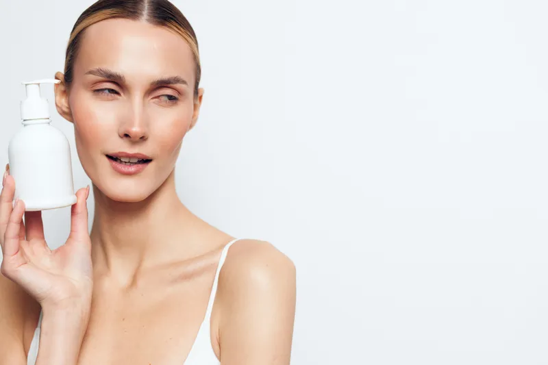 A focused woman with a white pump bottle demonstrates skincare with moisturizer, framed against a clean light backdrop, highlighting a daily beauty routine and self care.