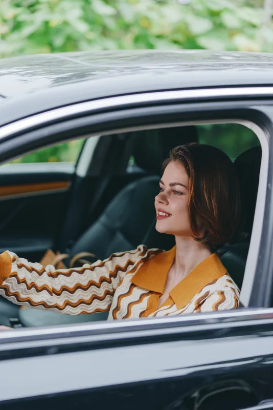 Portrait of a smiling woman seated in a car, relaxing with warm daylight and casual striped blouse.