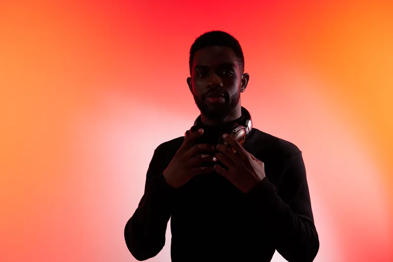 A stylish Black male DJ adjusting his headphones against a vibrant orange gradient background, expressing focus and creativity in a music scene