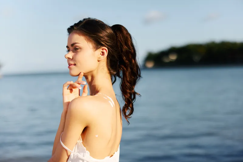 Smiling young woman with dark hair enjoying sunny day near calm blue sea water, wearing white dress in summer outdoors.