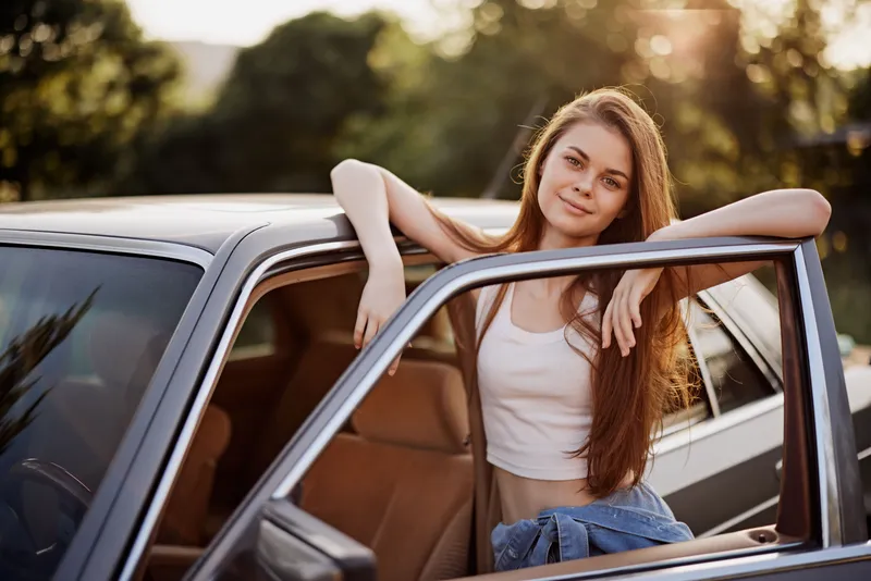 Young woman with long hair leaning on a car, smiling in the sunlight, wearing casual summer clothes and denim jacket, enjoying a relaxed moment outdoors.
