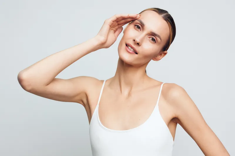 Woman portrait in a white tank top, touching the forehead with a gentle smile, soft studio lighting, clean neutral background, calm mood, simple casual beauty