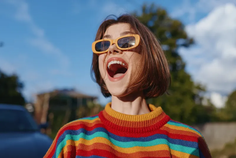 Young woman in her late 20s, smiling and laughing in a colorful rainbow sweater and yellow sunglasses with short hair, outdoor sunny suburban portrait conveying joyful lifestyle and fashion.