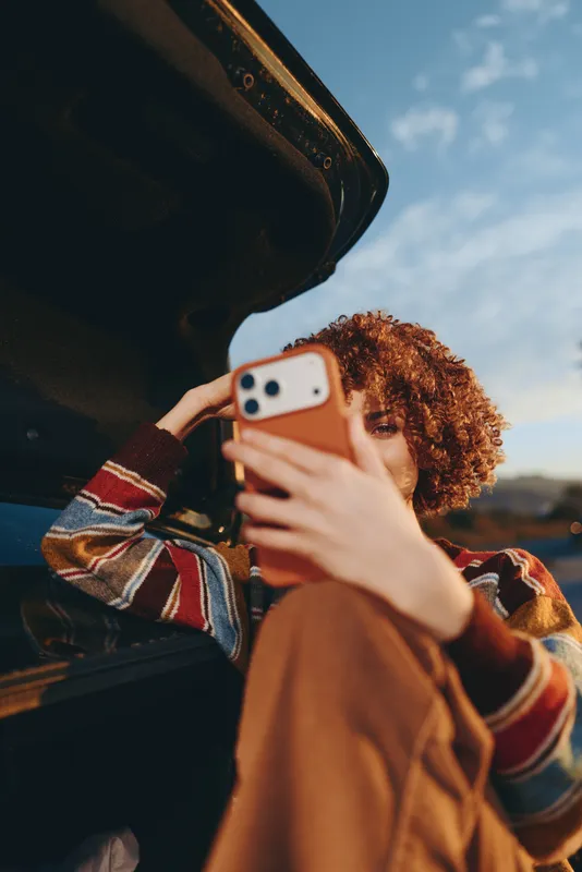 Woman lifestyle in a car, curly haired person holds smartphone for selfie, hand covering screen, warm daylight, outdoors view through window, casual travel and communication, mobile photography mood