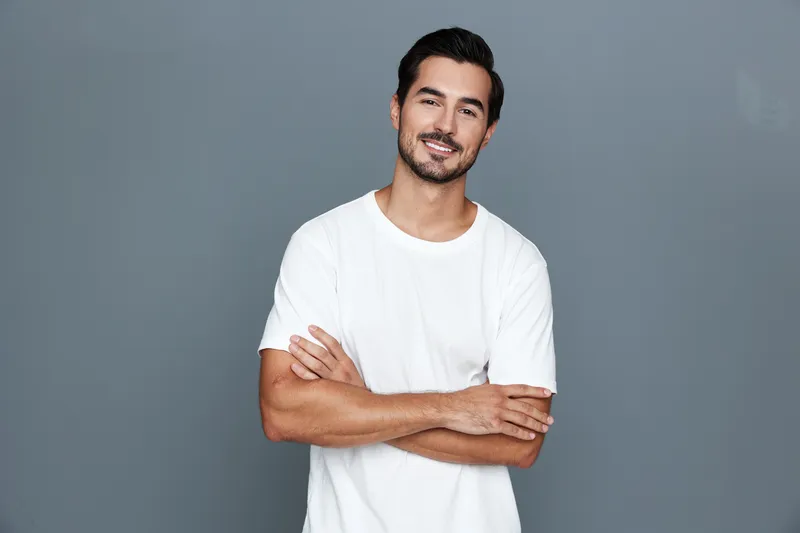 Smiling young man with crossed arms wearing a casual white t-shirt, looking confidently at the camera, isolated on a solid gray background. Person lifestyle concept.