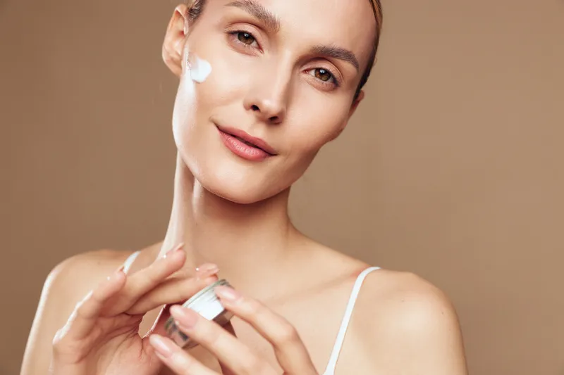 Young woman with clear skin applies moisturizing cream on her cheek while holding a small jar in a bright studio with a neutral background.