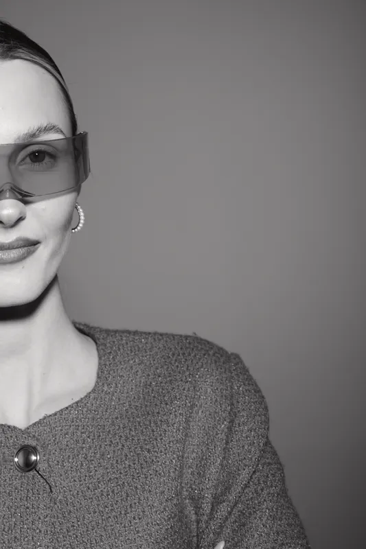 Portrait of a young white woman with fair skin wearing transparent sunglasses and a textured jacket. Studio shot with soft lighting and neutral grey background.