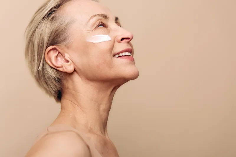 Mature woman with short hair applies white face cream on cheek, smiling and looking upwards. Skincare concept for aging skin, self-care, beauty routine, and natural wellness.