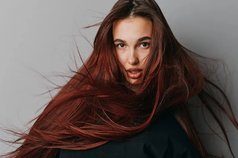 Woman with long red hair flowing around her face looking directly at camera with neutral expression on gray background. Portrait of female model with natural makeup and dynamic hair movement.