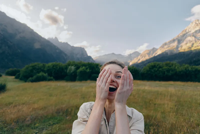 Young woman outdoors in a sunlit meadow laughs with her hands on her face, a carefree natural moment against distant mountains and a soft breeze.