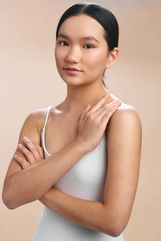 asian woman stands with arms crossed in a calm studio setting, confident and poised. soft lighting highlights natural complexion against a neutral background.