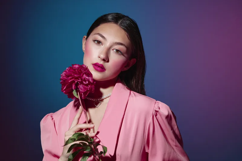 Young woman in pink outfit holds a dark red flower close to her face in a creative beauty fashion portrait with dramatic pink and blue lighting. Elegant and artistic mood.