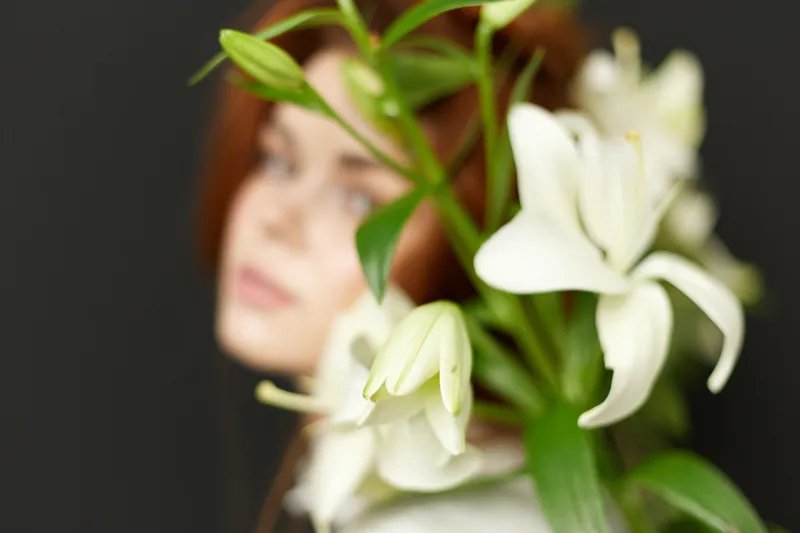 Close up of a young woman with flowing red hair, partially hidden behind a bouquet of white lilies, conveying serenity and elegance in a soft color palette