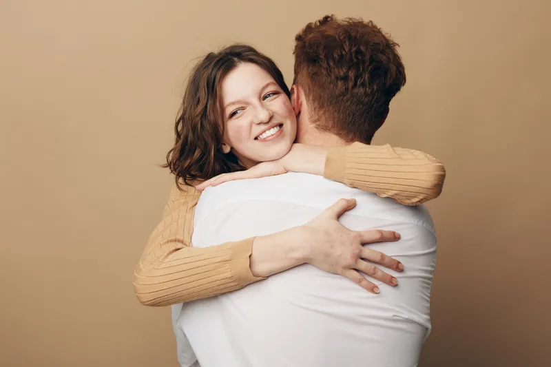 Happy young woman with brown hair hugs man wearing white shirt, smiling warmly with closed eyes. Intimate moment of affection, love, and connection between couple on beige background.