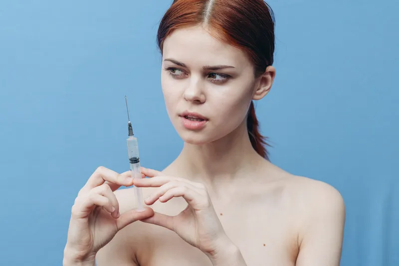 Женщина делает укол, её голые плечи и испуганное лицо на синем фоне