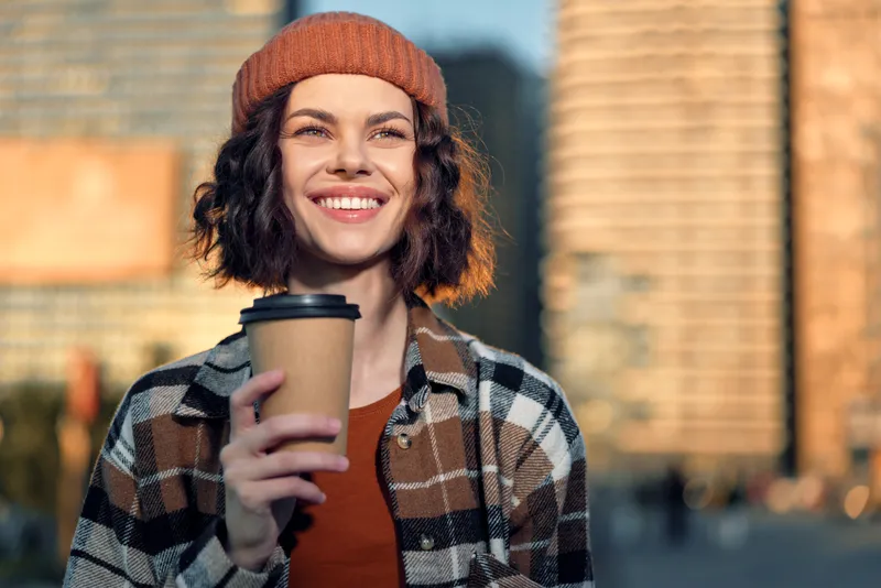 woman coffee smile urban beanie plaid, candid portrait of a young woman holding a takeaway cup in golden hour glow, authenticity and mindful living with emotional storytelling in warm city morning.