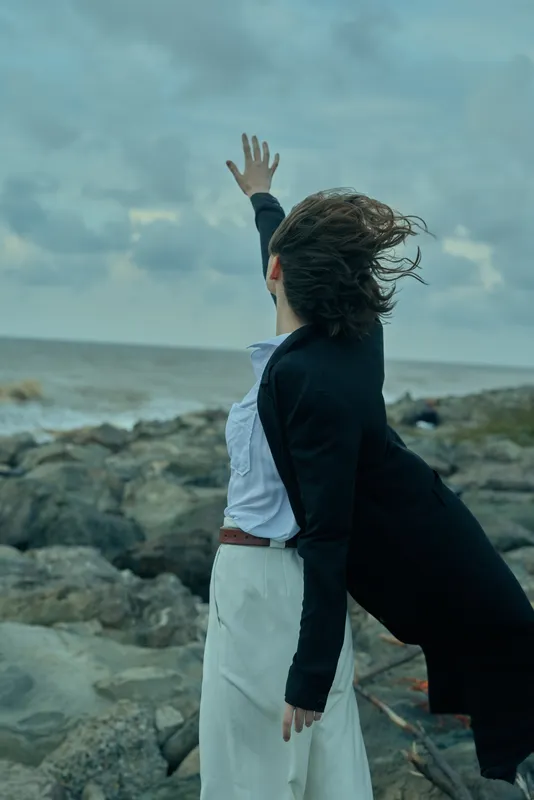 Woman on rocky shoreline reaches upward as wind sweeps her coat, ocean behind and cloudy sky above, capturing a dramatic outdoor moment of freedom, resilience and intense emotion in nature
