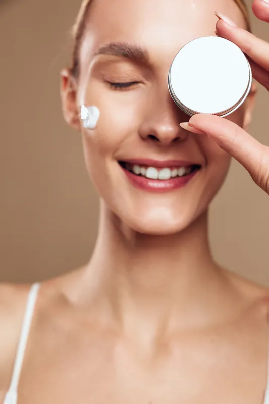Smiling woman with closed eyes applying cream on face holding cosmetic jar, clear skin, natural makeup, skincare routine, beige background, healthy beauty concept